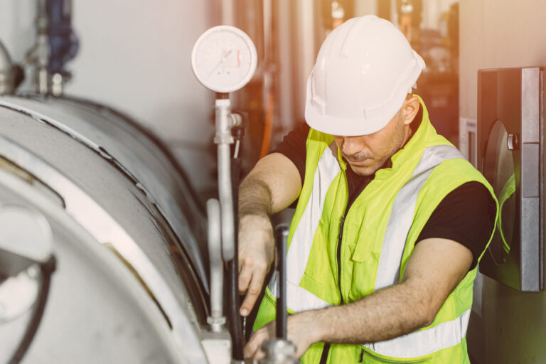 A factory service engineer i a white hard hat and green workers vest works on an industrial boiler at Combustion Technology